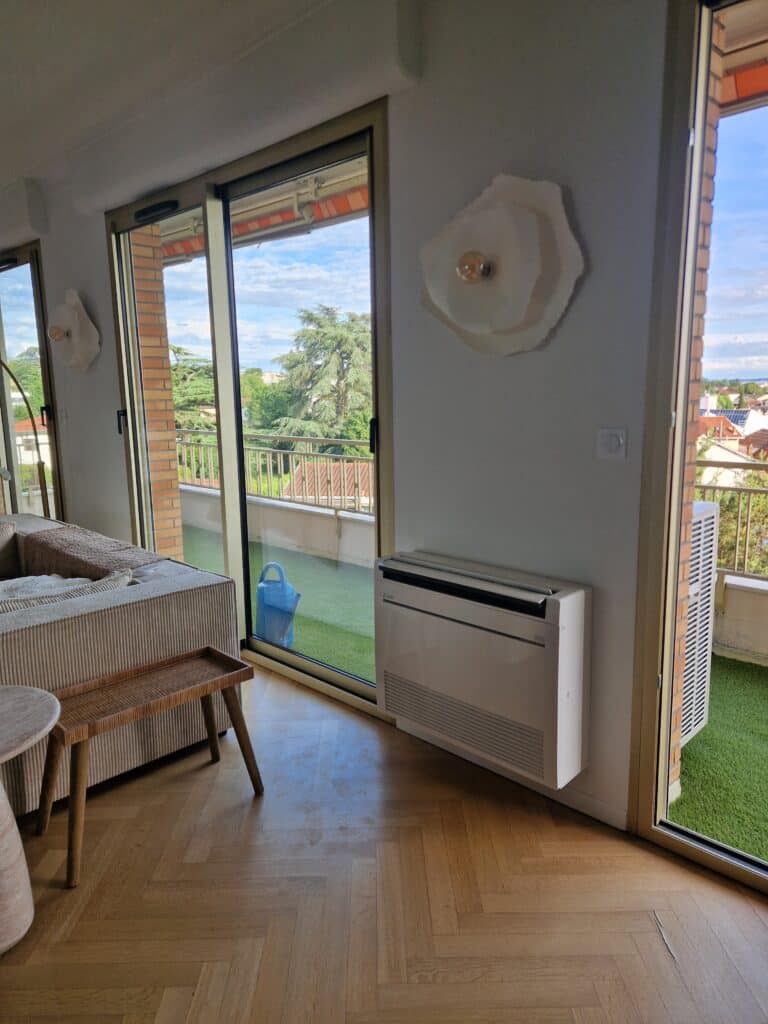 Salon moderne avec parquet, climatiseur mural et grande porte-fenêtre ouvrant sur un balcon en gazon synthétique.