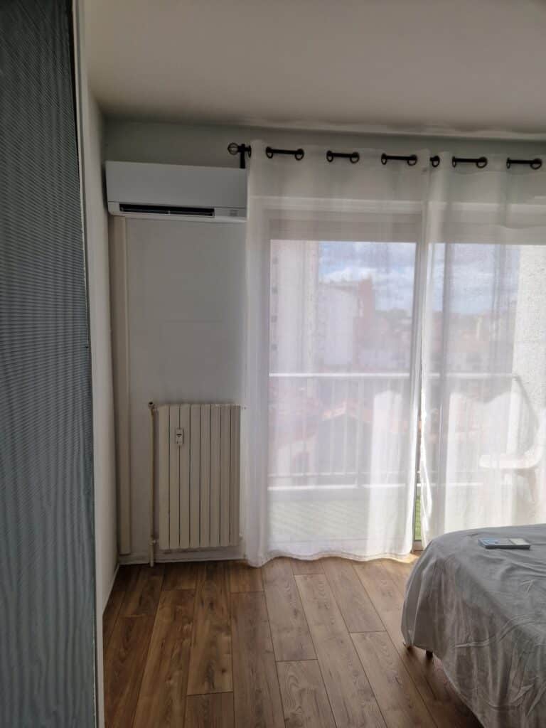 Climatiseur Mitsubishi et radiateur blanc installés près d'une baie vitrée avec rideaux blancs et parquet en bois.