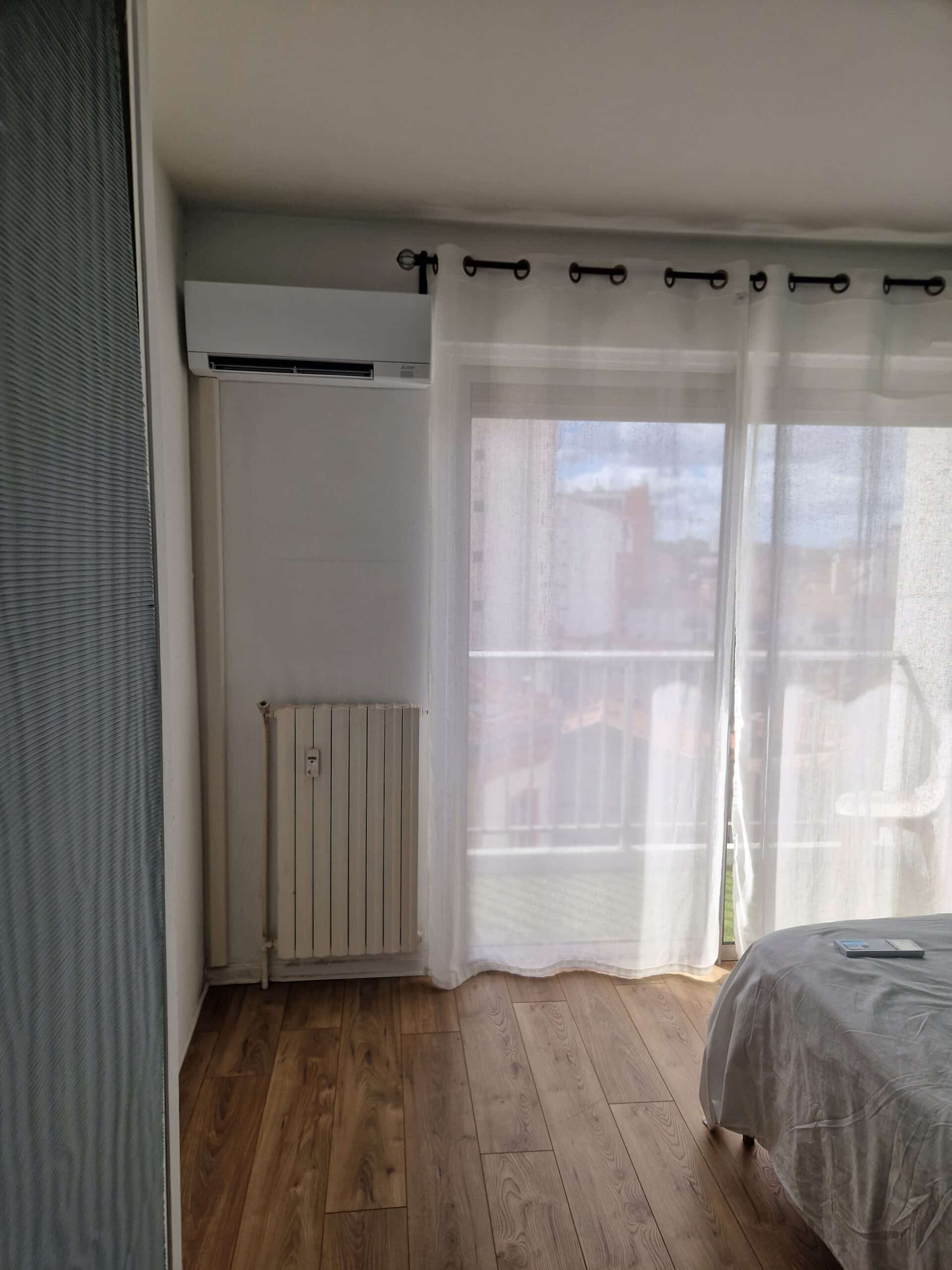 Installation Climatiseur et Radiateur dans une Chambre Climatiseur Mitsubishi et radiateur blanc installés près d'une baie vitrée avec rideaux blancs et parquet en bois.